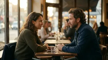 A man and woman sit at a cafe table, looking at each other. Coffee and water are on the table in warm light