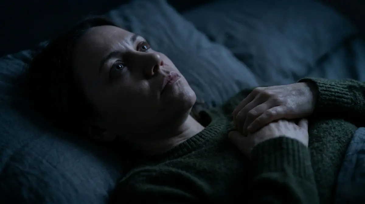 A person lying awake in a darkened bedroom with hand on chest, representing the insomnia and physical discomfort linked to trauma-related nervous system dysregulation.