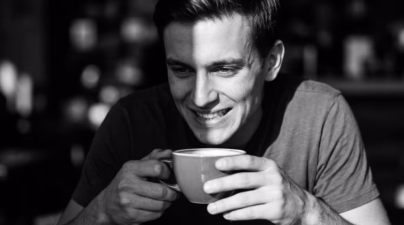 A young man holding a coffee cup with a tense expression and forced smile in a cafe, demonstrating the chronic fawning autonomic response common in adult family scapegoat characteristics.